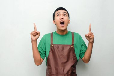 Yeşil gömlekli ve kahverengi önlüklü genç adam iki işaret parmağı ve şaşırmış bir ifadeyle yukarı bakıyor. İlgi, keşif ya da beklenmedik an kavramı