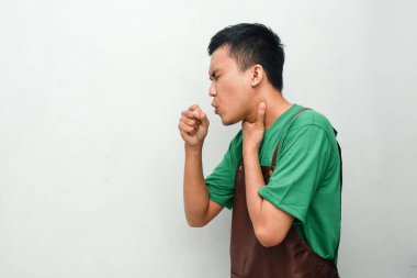 Yeşil tişörtlü ve kahverengi önlüklü gülümseyen Asyalı genç adam, dost canlısı bir ifadeyle öksürük jesti yapıyor. Sağlık kavramı, farkındalık, ya da tesadüfi an kavramı