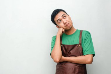 Yeşil gömlekli ve kahverengi önlüklü genç adam kafası karışık bir ifadeyle düşünceli bir hareket yapıyor. Karar verme, sorun çözme veya belirsizlik kavramı