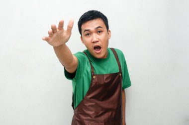 Yeşil tişörtlü ve kahverengi önlüklü gülümseyen Asyalı genç paniklemiş bir ifadeyle korkmuş bir jest yapıyor. Hizmet bağlamında sürpriz, stres veya beklenmedik durum kavramı
