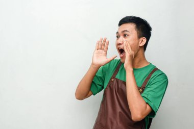 Yeşil tişörtlü ve kahverengi önlüklü gülümseyen Asyalı genç coşkulu bir ifadeyle bağırarak bir jest yapıyor. Heyecan, motivasyon ya da duyuru kavramı