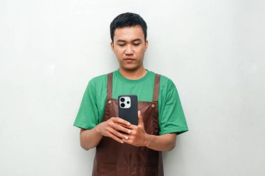 Yeşil tişörtlü ve kahverengi önlüklü genç adam sakin bir ifadeyle telefon kullanıyor. Dijital aktivite kavramı, rahatlama, ya da günlük iş anı