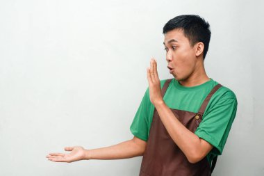 Yeşil gömlekli ve kahverengi önlüklü genç adam yan tarafı işaret ediyor, sanki bir ürünü sunuyormuş gibi, şaşırmış bir ifadeyle. Keşif kavramı, promosyon veya özellikli öge