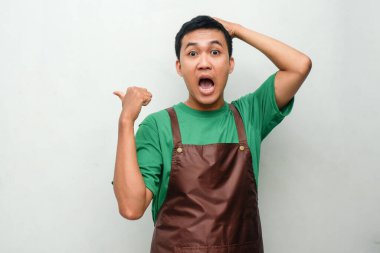 Yeşil gömlekli ve kahverengi önlüklü genç adam şaşırmış bir ifadeyle başını tutarken yan tarafı işaret ediyor. Şok kavramı, farkındalık, ya da beklenmedik an