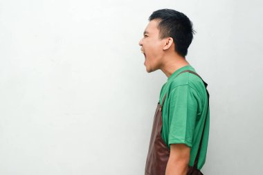 Yeşil gömlekli ve kahverengi önlüklü genç adam bağırarak jest yapıyor.