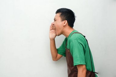 Yeşil gömlekli ve kahverengi önlüklü genç adam bağırarak jest yapıyor.