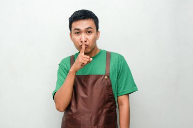 Yeşil gömlekli ve kahverengi önlüklü genç adam sakin bir ifadeyle ağzını kapatıyor ya da sır saklıyor.