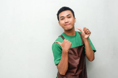 Yeşil tişörtlü ve kahverengi önlüklü genç bir adam. Dostça bir yüz ifadesi ve iki başparmağıyla yüzünü gösteren bir gülümseme.