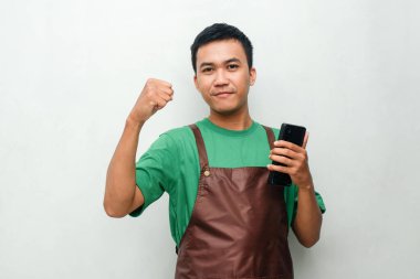 Yeşil tişörtlü ve kahverengi önlüklü genç adam telefonu tutarken dostça bir yüz ifadesiyle yumruk hareketi yapıyor.