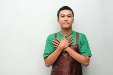 Yeşil tişörtlü, kahverengi önlüklü, güler yüzlü genç adam göğsünü tutuyor.