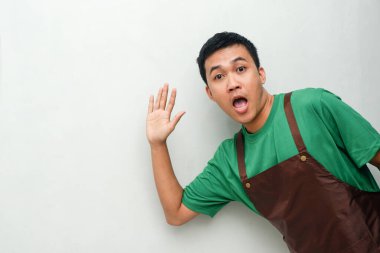 Yeşil gömlekli ve kahverengi önlüklü genç adam karşılama hareketi yapıyor.