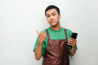 Yeşil tişörtlü ve kahverengi önlüklü genç adam telefonu tutarken yüzünde dostane bir ifadeyle baş parmağıyla işaret ediyor.