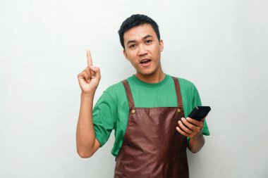 Yeşil tişörtlü ve kahverengi önlüklü genç adam işaret parmağıyla yukarı işaret ediyor.