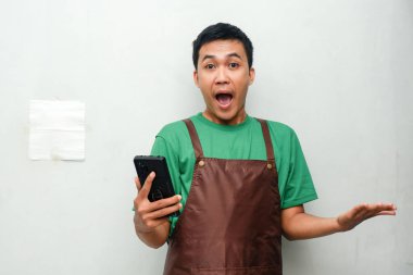 Yeşil tişörtlü ve kahverengi önlüklü genç adam şaşırmış bir ifade sergiliyor ve telefonu tutuyor.