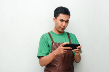 Yeşil gömlekli ve kahverengi önlüklü genç adam komik bir ifadeyle telefonu tutuyor ya da oyun oynuyor.