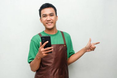 Yeşil tişörtlü ve kahverengi önlüklü genç adam. Dostane bir ifade takınıyor ve telefonu tutuyor.