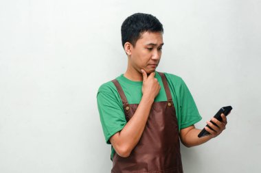 Yeşil tişörtlü ve kahverengi önlüklü genç adam telefonuna bakıyor.