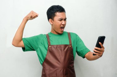 Yeşil tişörtlü ve kahverengi önlüklü genç adam telefonu tutuyor ve zaferi kutlamak için jest yapıyor.