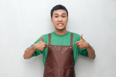 Yeşil gömlekli ve kahverengi önlüklü gülümseyen genç adam iki parmağını kaldırıyor. Neşeli ve dostça bir ifade, barista veya hizmet sektörü rolleri için mükemmel.