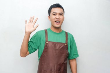 Yeşil gömlekli ve kahverengi önlüklü gülümseyen genç adam kameraya el sallıyor. Neşeli ve dostça bir ifade, hizmet ve konukseverlik için mükemmel.