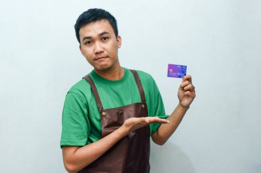 Yeşil tişörtlü ve kahverengi önlüklü genç adam kredi kartını gösteriyor.