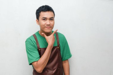 Yeşil gömlekli ve kahverengi önlüklü gülümseyen genç adam kameranın önünde poz veriyor. Neşeli ve dostane ifade, hizmet ve konukseverlik rolleri için ideal.