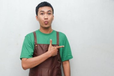 Yeşil gömlekli, kahverengi önlüklü, solu gösteren gülümseyen genç adam. Keyifli ve dostça bir ifade, hizmet veya tanıtım amaçlarına uygun.