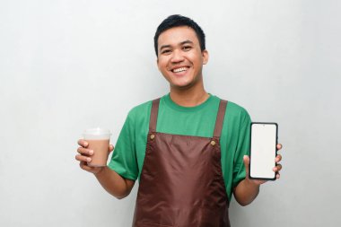 Yeşil tişörtlü ve kahverengi önlüklü gülümseyen Asyalı genç adam dostça ve coşkulu bir ifadeyle başparmak kaldırıyor. Servis, kafe veya tanıtım temaları için mükemmel