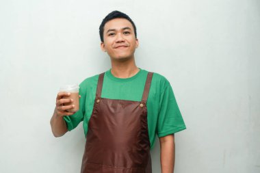 Yeşil tişörtlü ve kahverengi önlüklü gülümseyen Asyalı genç adam dostça ve coşkulu bir ifadeyle başparmak kaldırıyor. Servis, kafe veya tanıtım temaları için mükemmel