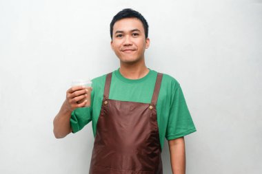 Yeşil tişörtlü ve kahverengi önlüklü gülümseyen Asyalı genç adam dostça ve coşkulu bir ifadeyle başparmak kaldırıyor. Servis, kafe veya tanıtım temaları için mükemmel