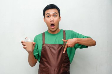 Yeşil tişörtlü ve kahverengi önlüklü gülümseyen Asyalı genç adam dostça ve coşkulu bir ifadeyle başparmak kaldırıyor. Servis, kafe veya tanıtım temaları için mükemmel