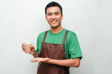 Yeşil tişörtlü ve kahverengi önlüklü gülümseyen Asyalı genç adam dostça ve coşkulu bir ifadeyle başparmak kaldırıyor. Servis, kafe veya tanıtım temaları için mükemmel