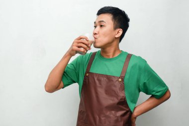 Yeşil tişörtlü ve kahverengi önlüklü gülümseyen Asyalı genç adam dostça ve coşkulu bir ifadeyle başparmak kaldırıyor. Servis, kafe veya tanıtım temaları için mükemmel