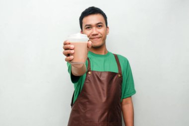 Yeşil tişörtlü ve kahverengi önlüklü gülümseyen Asyalı genç adam dostça ve coşkulu bir ifadeyle başparmak kaldırıyor. Servis, kafe veya tanıtım temaları için mükemmel