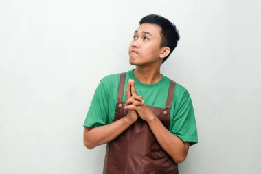 Yeşil tişörtlü ve kahverengi önlüklü gülümseyen Asyalı genç adam dostça ve coşkulu bir ifadeyle başparmak kaldırıyor. Servis, kafe veya tanıtım temaları için mükemmel