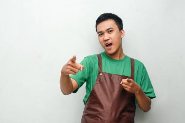 Yeşil tişörtlü ve kahverengi önlüklü gülümseyen genç adam her iki işaret parmağıyla, dostane ve coşkulu bir ifadeyle ileri bakıyor. Promosyon, hizmet ve hoş geldin teması için mükemmel.