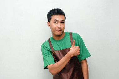 Yeşil tişörtlü ve kahverengi önlüklü gülümseyen Asyalı genç adam dostça ve coşkulu bir ifadeyle başparmak kaldırıyor. Servis, kafe veya tanıtım temaları için mükemmel