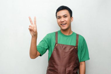 Yeşil tişörtlü ve kahverengi önlüklü gülümseyen Asyalı genç adam dostça ve coşkulu bir ifadeyle başparmak kaldırıyor. Servis, kafe veya tanıtım temaları için mükemmel