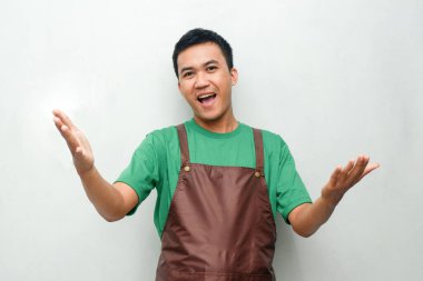 Yeşil tişörtlü ve kahverengi önlüklü gülümseyen Asyalı genç adam dostça ve coşkulu bir ifadeyle başparmak kaldırıyor. Servis, kafe veya tanıtım temaları için mükemmel