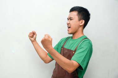 Yeşil gömlekli ve kahverengi önlüklü gülümseyen genç adam dostça ve coşkulu bir ifadeyle zafer kutlaması yapıyor. Başarı, hizmet veya tanıtım temaları için mükemmel