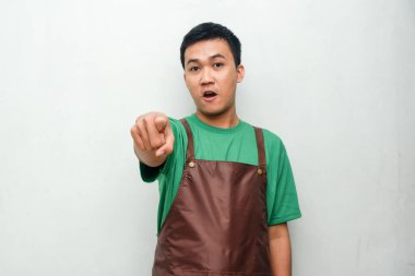 Yeşil tişörtlü ve kahverengi önlüklü gülümseyen Asyalı genç adam işaret parmağıyla, dostane ve coşkulu bir ifadeyle ileri doğru işaret ediyor. Promosyon veya servis temaları için harika
