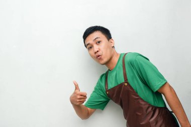 Yeşil tişörtlü ve kahverengi önlüklü gülümseyen Asyalı genç adam dostça ve coşkulu bir ifadeyle başparmak kaldırıyor. Servis, kafe veya tanıtım temaları için mükemmel