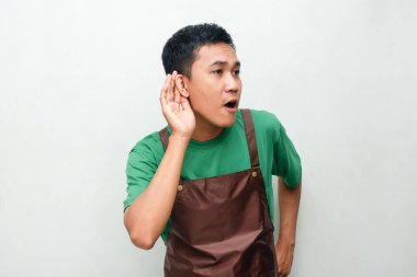 Yeşil tişörtlü ve kahverengi önlüklü gülümseyen Asyalı genç adam arkadaş canlısı ve coşkulu bir ifadeyle dinleyici bir jest yapıyor. Müşteri hizmetleri veya iletişim temaları için uygundur