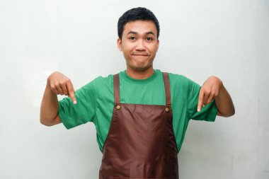 Yeşil tişörtlü ve kahverengi önlüklü gülümseyen Asyalı genç adam her iki işaret parmağıyla aşağıya doğru bakıyor, arkadaş canlısı ve coşkulu bir ifadeyle. Promosyon, rehber veya hizmet temaları için ideal