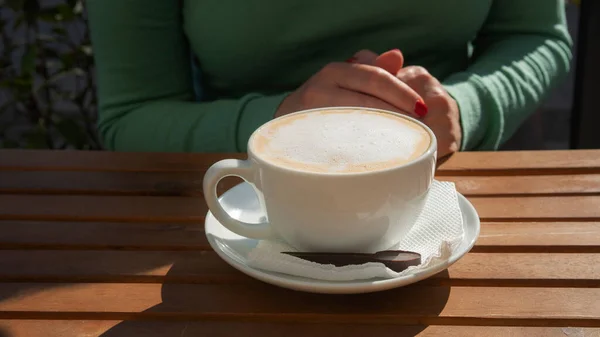 Girl at the table in a cafe with a cup of cappuccino. Delicious drink with appetizing milk foam. Cup of coffee close-up. Breakfast outdoors.