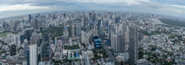 Bangkok, Tayland 'ın panoramik manzarası modern gökdelenler, yeşil alanlar ve dolambaçlı caddelerle dolu geniş bir şehir manzarası sergiliyor. Bulutlu gökyüzü hareketli ve dinamik şehir manzarasına derinlik katıyor..