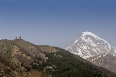 Gürcistan 'daki Kazbek Dağı' nın nefes kesici manzarası, açık mavi gökyüzünün altında görkemli bir şekilde yükselen karlı zirvesi. Tarihi bir kilise yeşil bir tepenin üzerinde, ormanlarla çevrili, doğa ve kültürü harmanlayan.