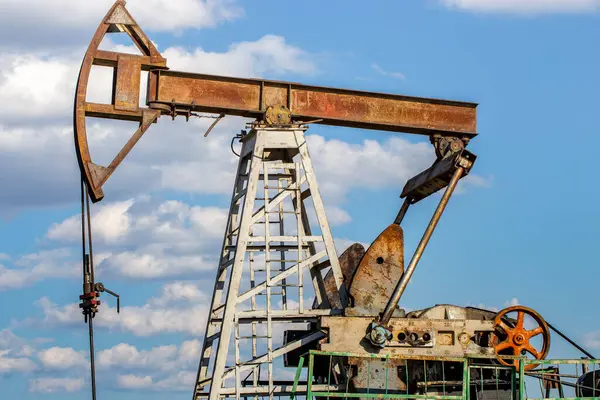 Petrol çıkarmak için kullanılan yıpranmış bir petrol pompacısı dağınık bulutlu parlak mavi gökyüzüne karşı. Metal yapı paslıdır, görünür dişliler, kirişler ve enerji üretimini sembolize eden turuncu bir tekerlekle..