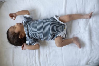 Bir bebek kolları açık beyaz bir çarşafın üzerinde yatıyor. Bebeğin üzerinde siyah beyaz çizgili bir gömlek var.