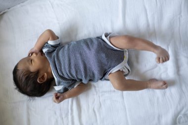 Bir bebek kolları açık beyaz bir çarşafın üzerinde yatıyor. Bebeğin üzerinde siyah beyaz çizgili bir gömlek var.